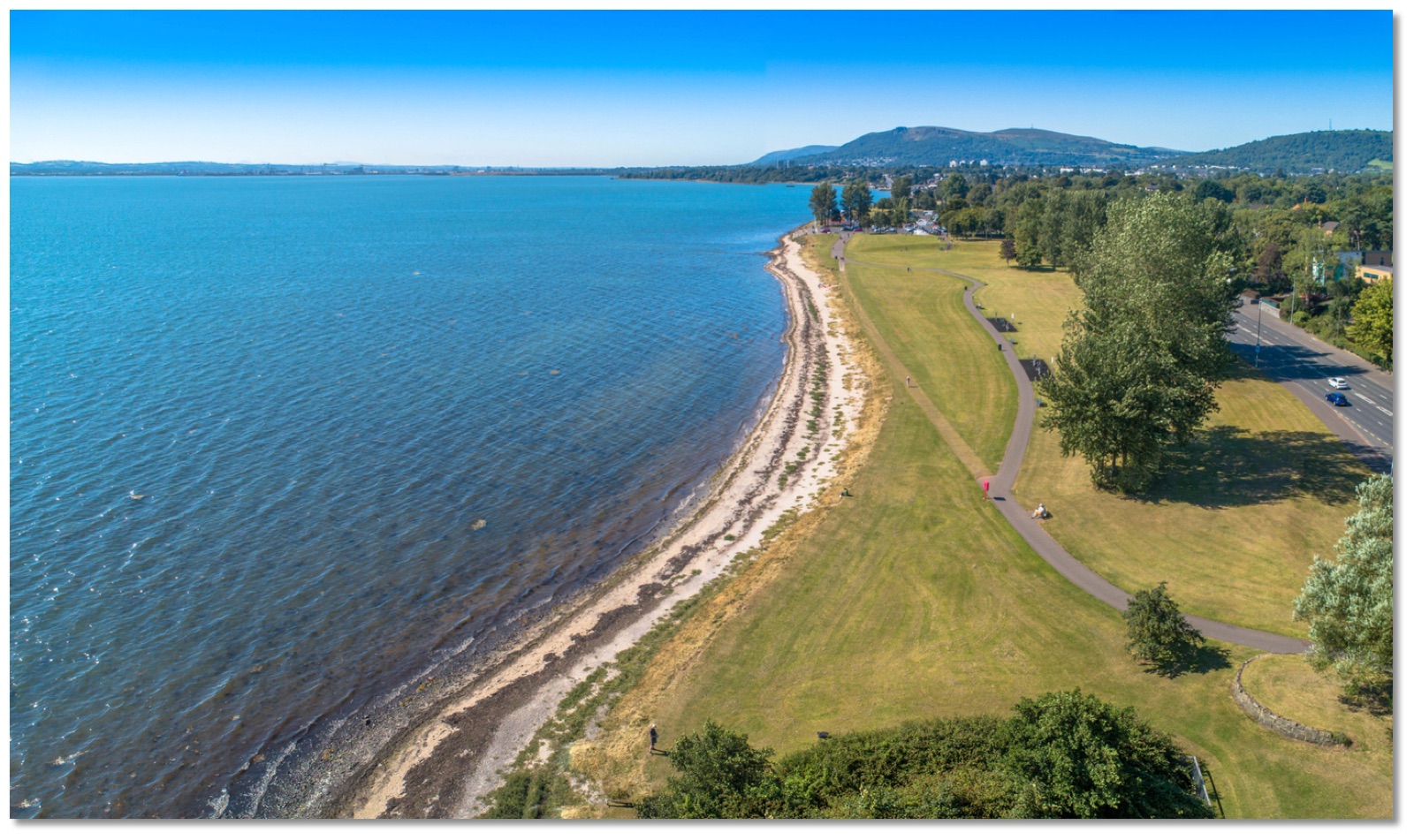 Aerial view along the curving shoreline at Jordanstown with turquoise water, a shingle beach and the green park strip