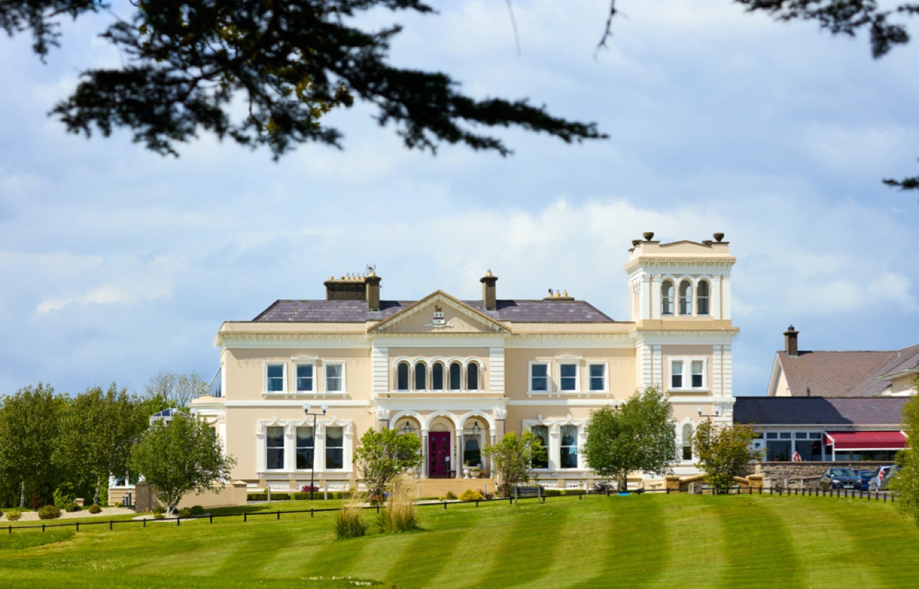 The cream front of the manor hotel seen across a sweeping lawn under a blue sky