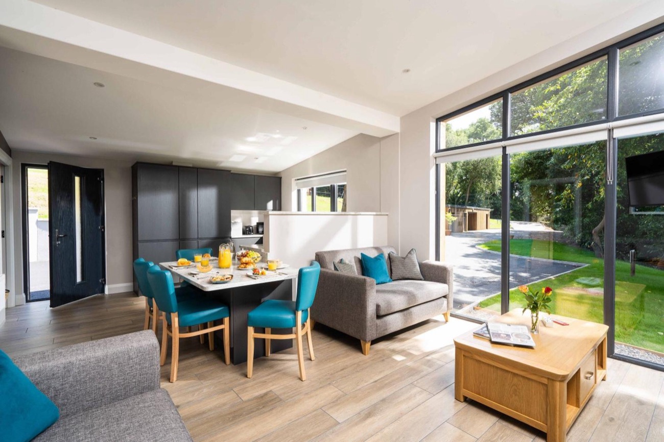 A bright open-plan chalet interior with a kitchen, dining table and sofa opening onto floor-to-ceiling windows and the garden
