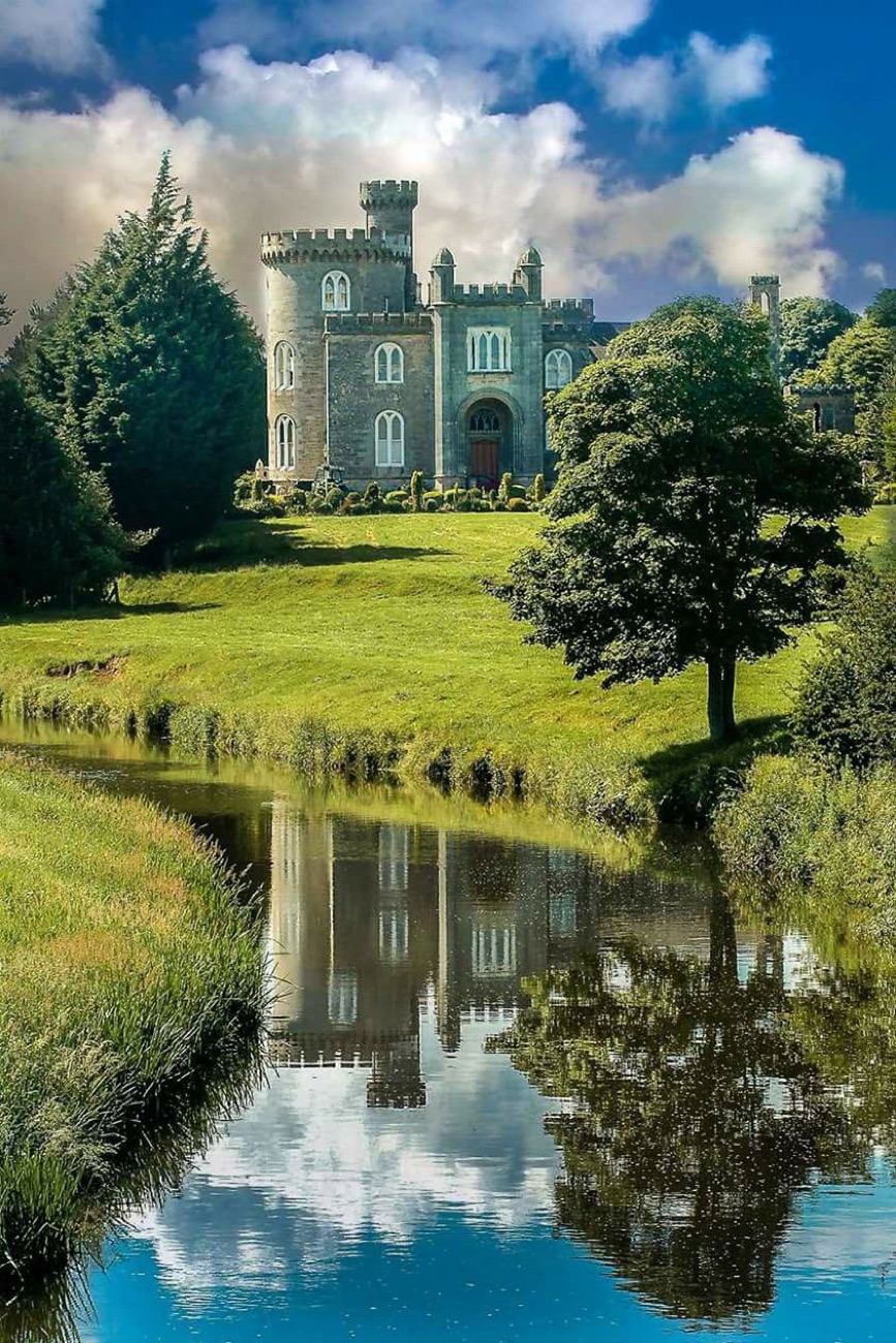 Killymoon Castle reflected in the calm water of the Ballinderry River, framed by a grassy bank and a single tree