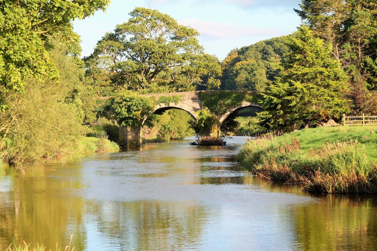 The Ballinderry river running under an old stone arch bridge in the Killymoon demesne, with trees on both banks in golden light
