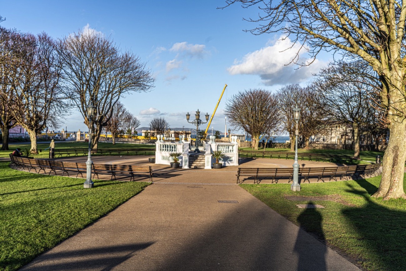 A tree-lined path in the park leading towards the promenade with the harbour crane beyond