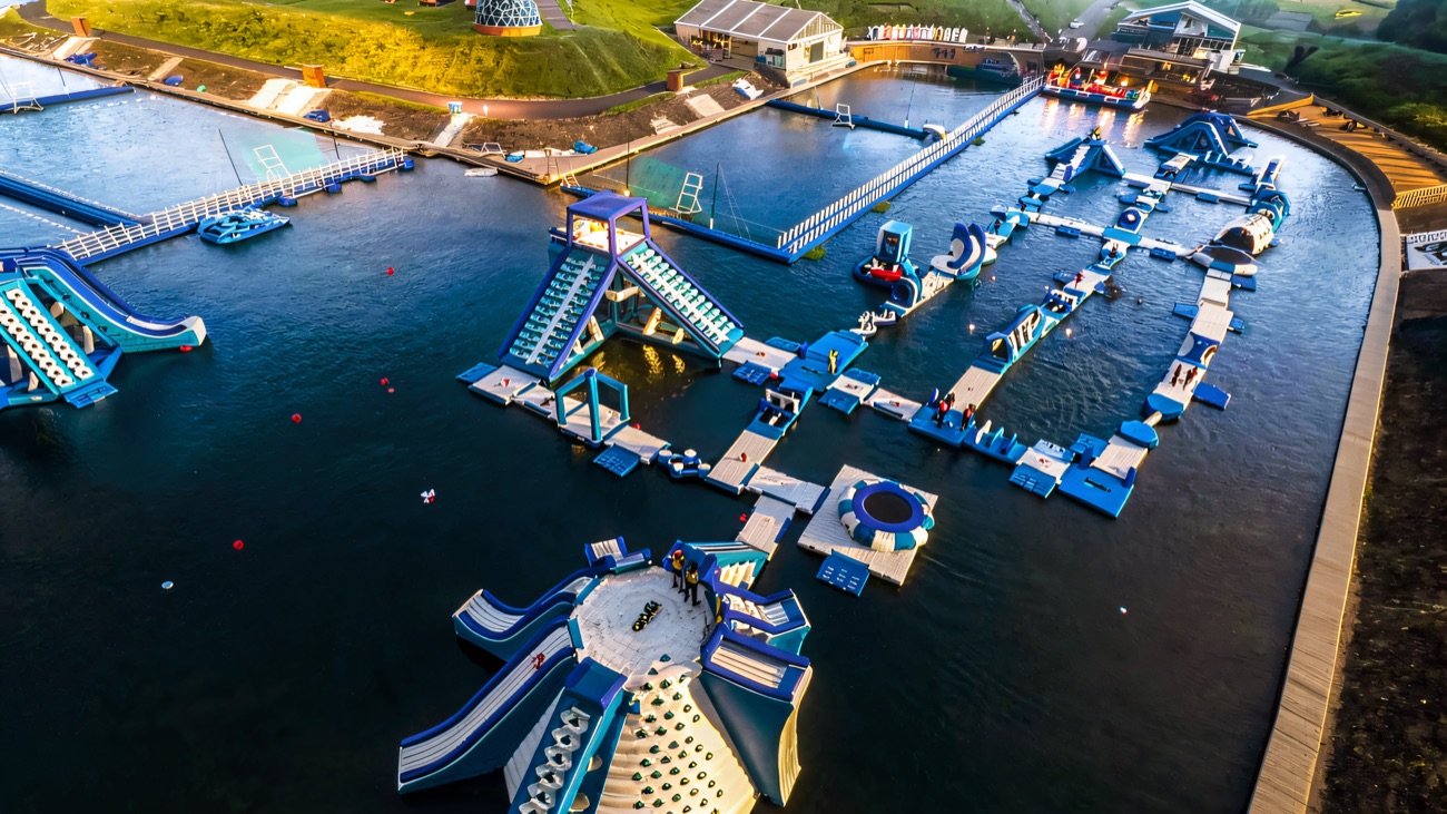 Aerial view of the blue-and-white floating obstacle course with the central hydro island, jump platforms and the resort buildings behind