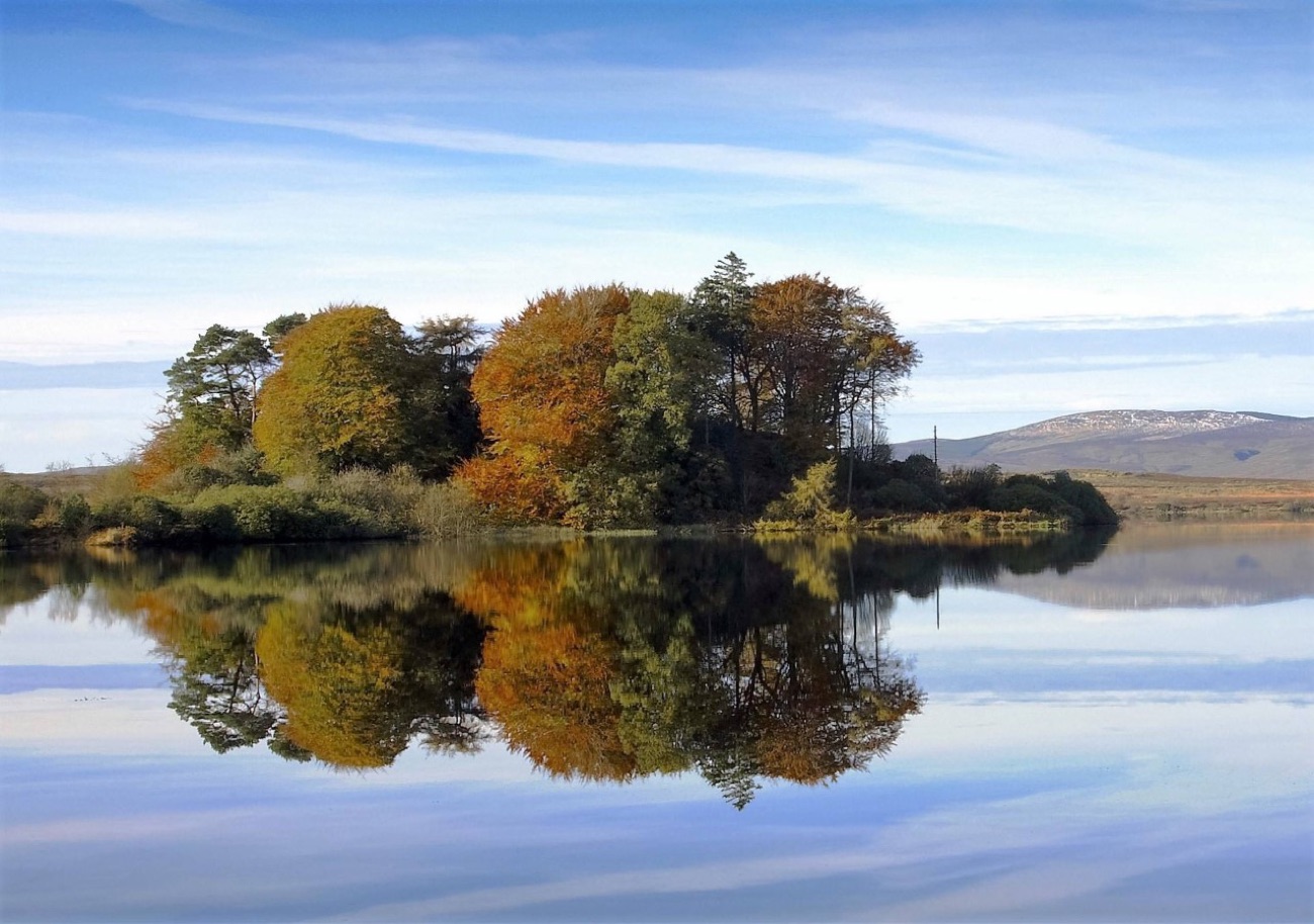 The wooded island at Lough Fea in autumn colour, perfectly reflected in still water with snow on the far hills