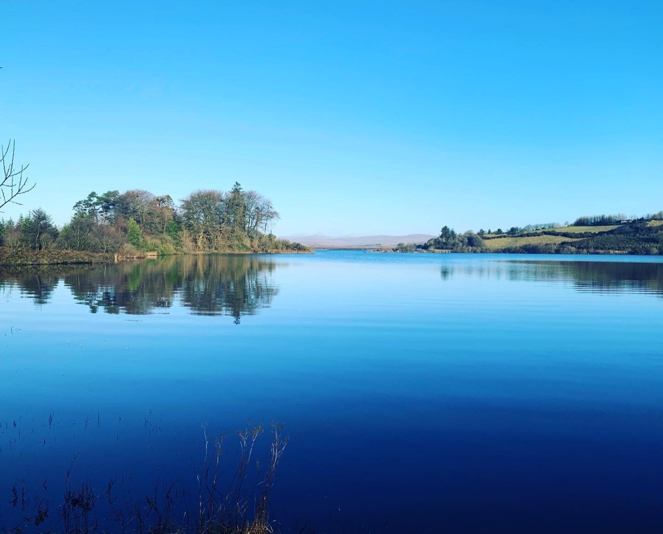 Lough Fea on a calm blue day with the wooded island reflected and the hills beyond