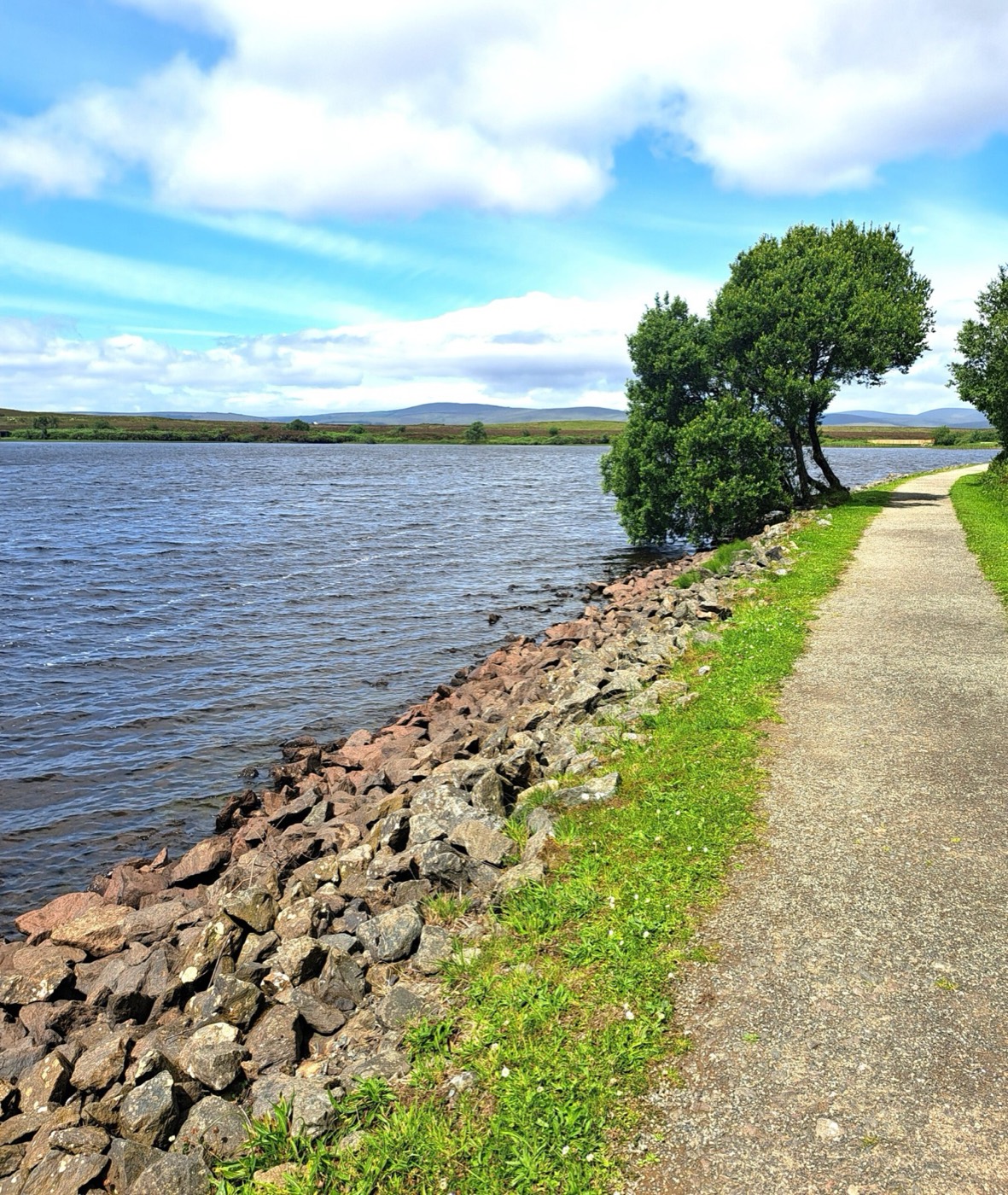 The gravel lakeside path at Lough Fea running along the stone shore with the Sperrin hills behind