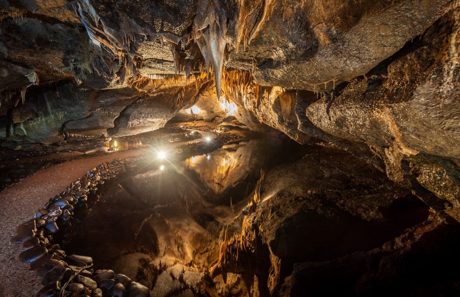 A lit underground cave chamber with a still, mirror-like pool reflecting stalactites, and a lantern-lit walkway