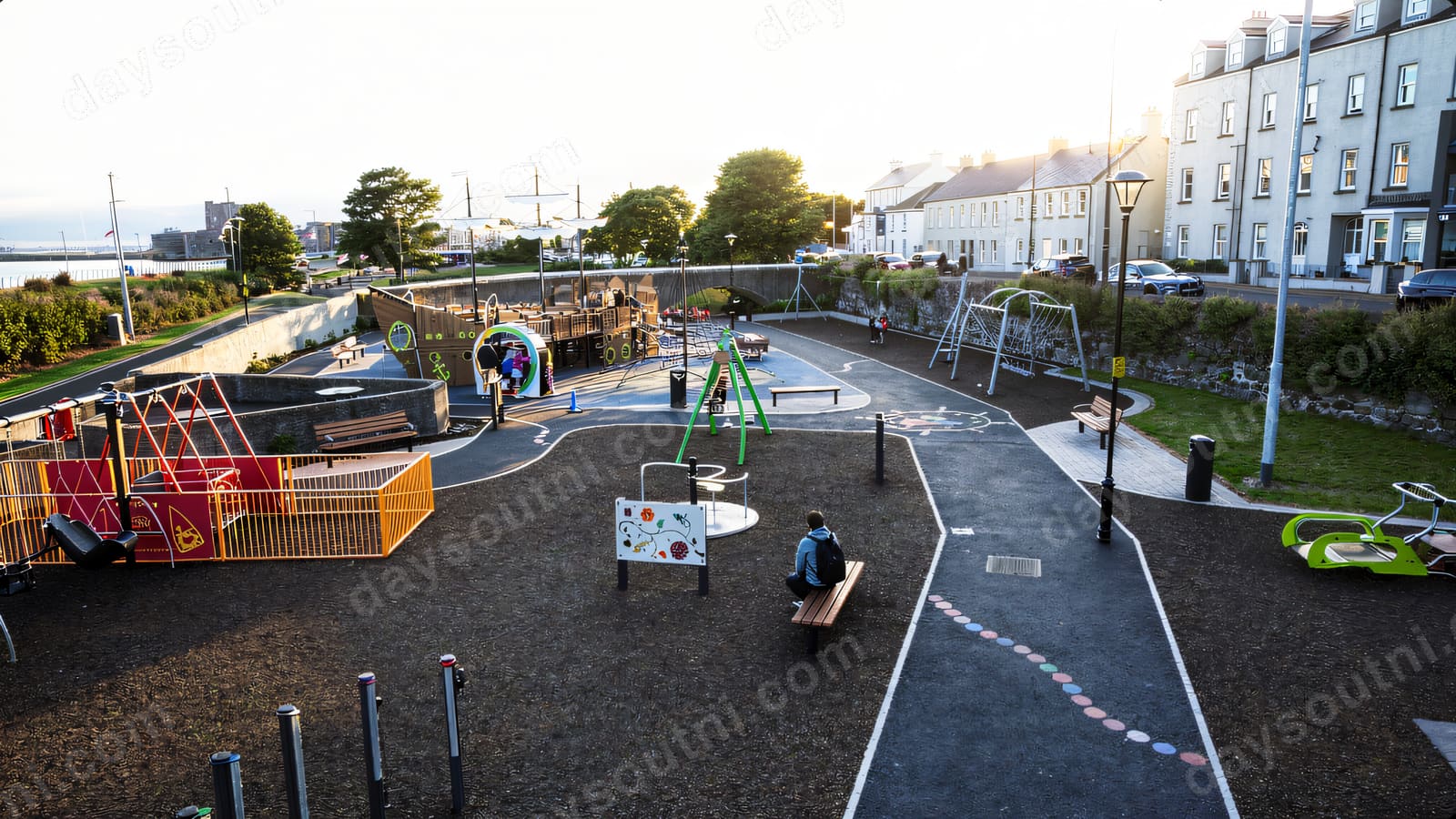 Swings and a roundabout on tarmac paths with the seafront street and houses behind