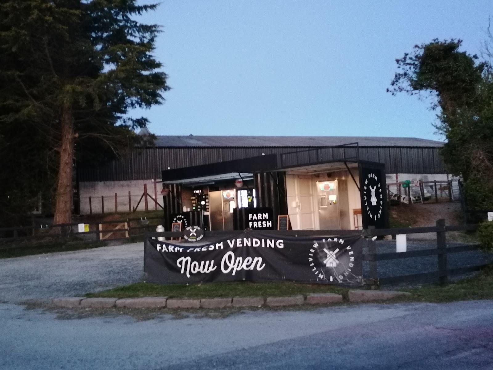 The Millview Farm Vending shed lit up in the evening with a Farm Fresh Vending Now Open banner along the front