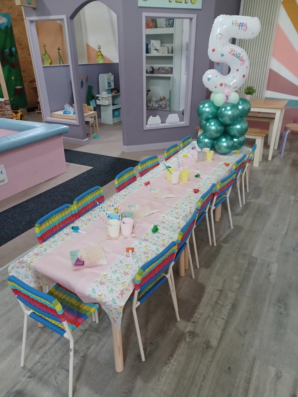 A birthday party table set up within the Mini Ville mini-town, with a pastel cloth, balloons and the pretend shops behind