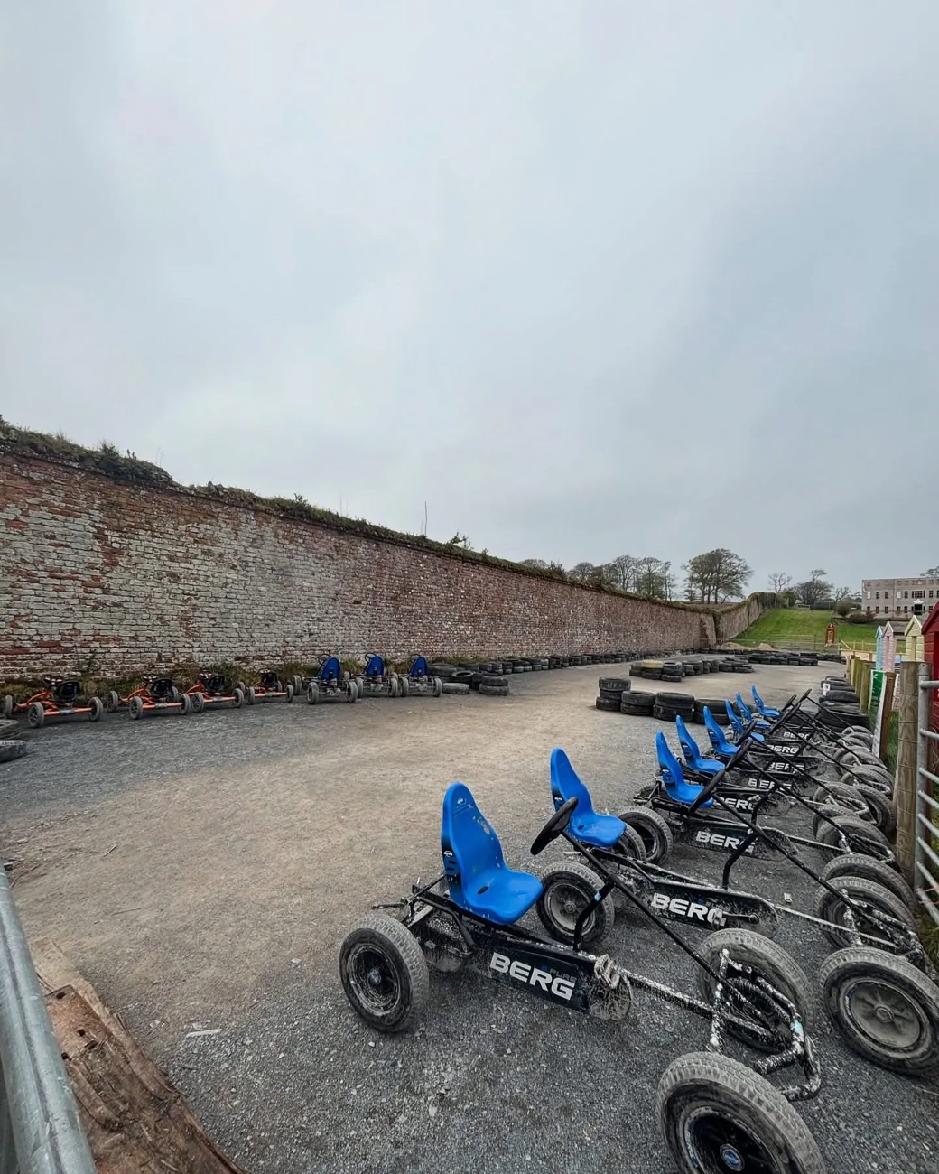 A row of pedal go-karts on the gravel track beside the tall old brick wall