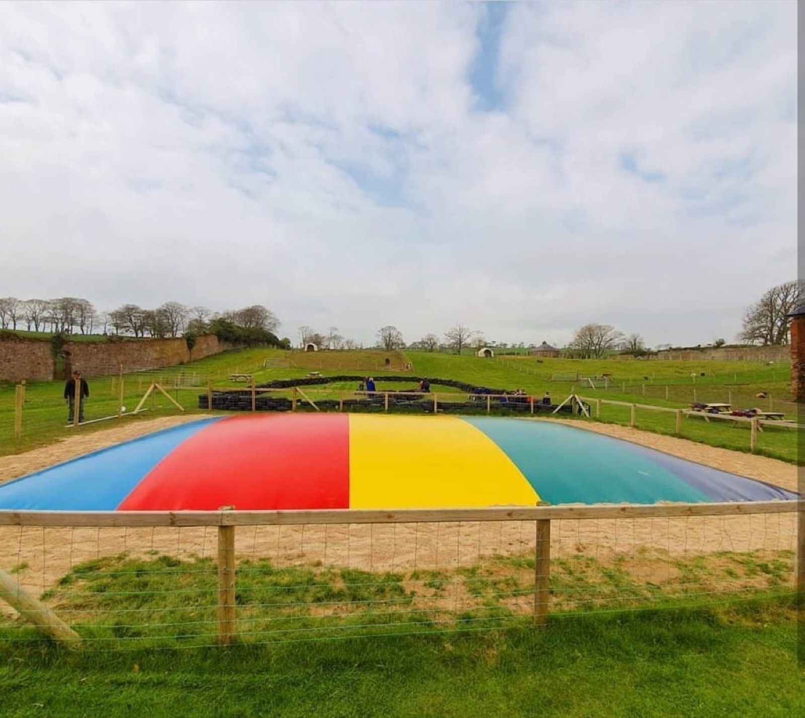 The big rainbow jumping pillow set in sand at Mountpanther Farm Park, with rolling green hills and the deer field behind