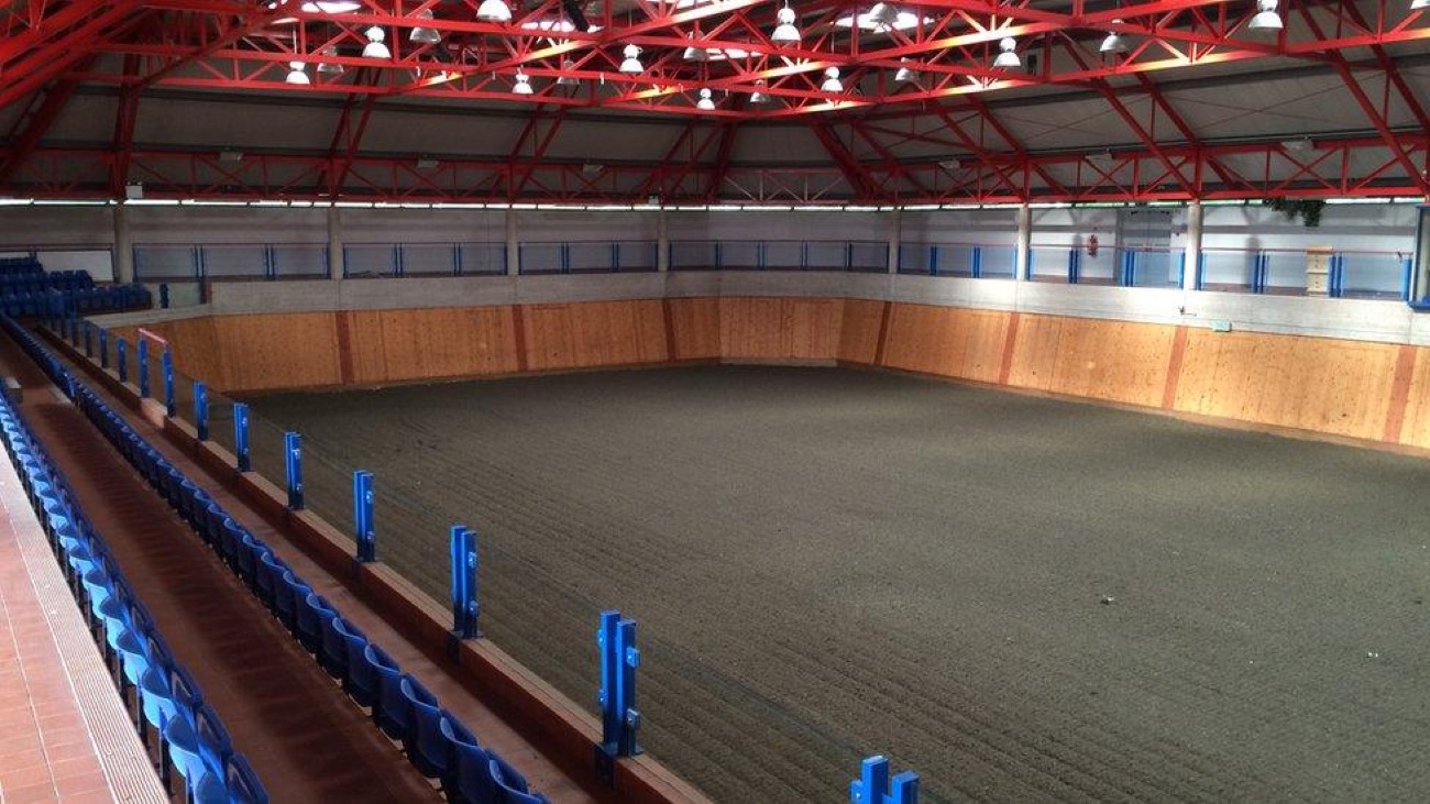 The large indoor arena at Necarne equestrian centre, red steel roof and tiered blue spectator seating around a sand surface