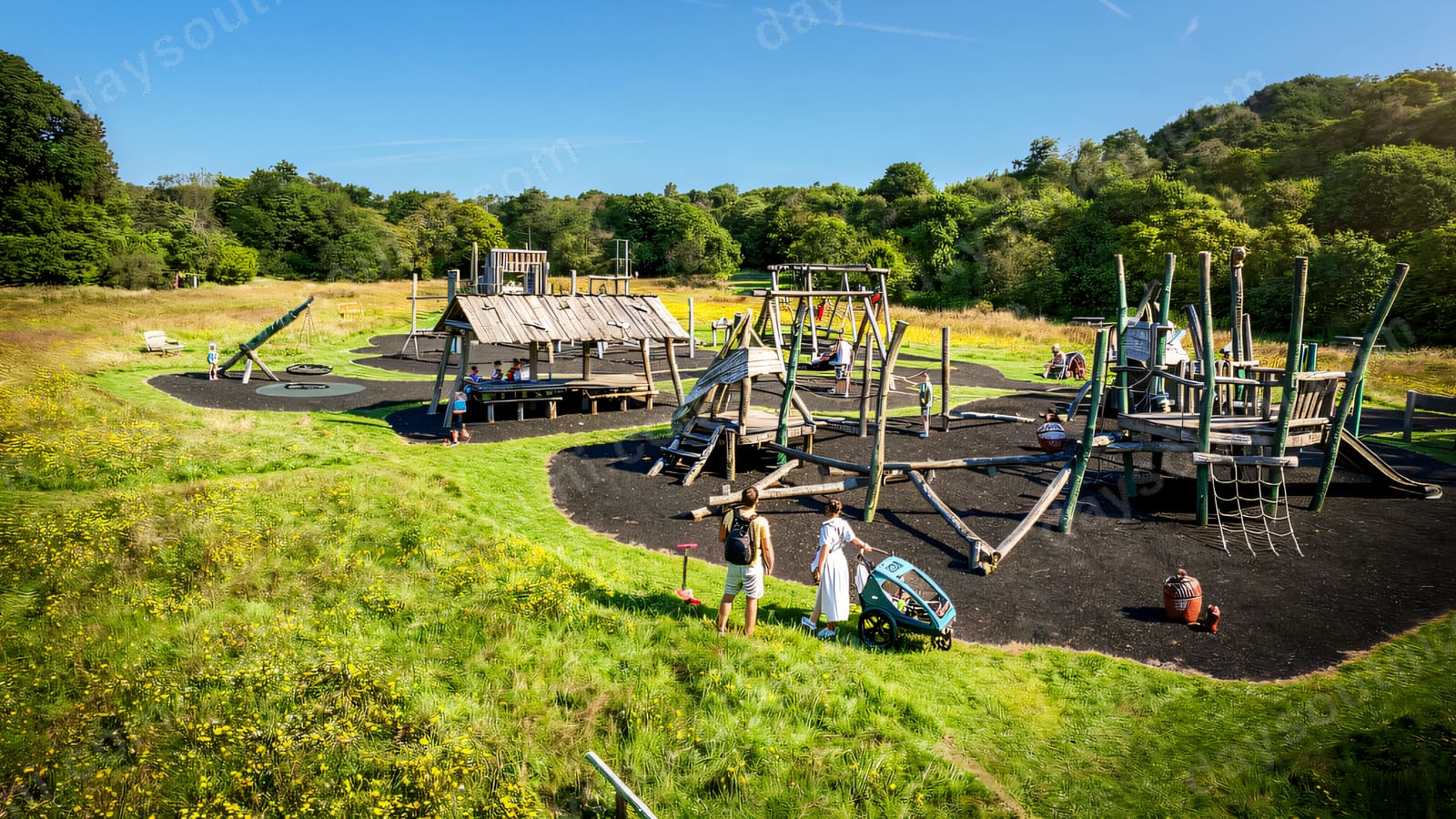 A long timber and steel adventure playground set in rough grass with woodland behind.