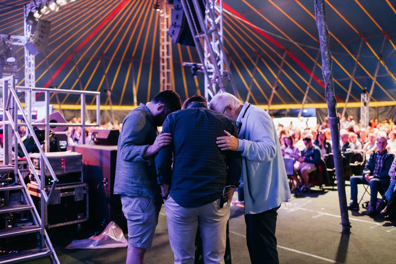 Three people praying together at the front of the New Horizon main tent, with the crowd seated behind