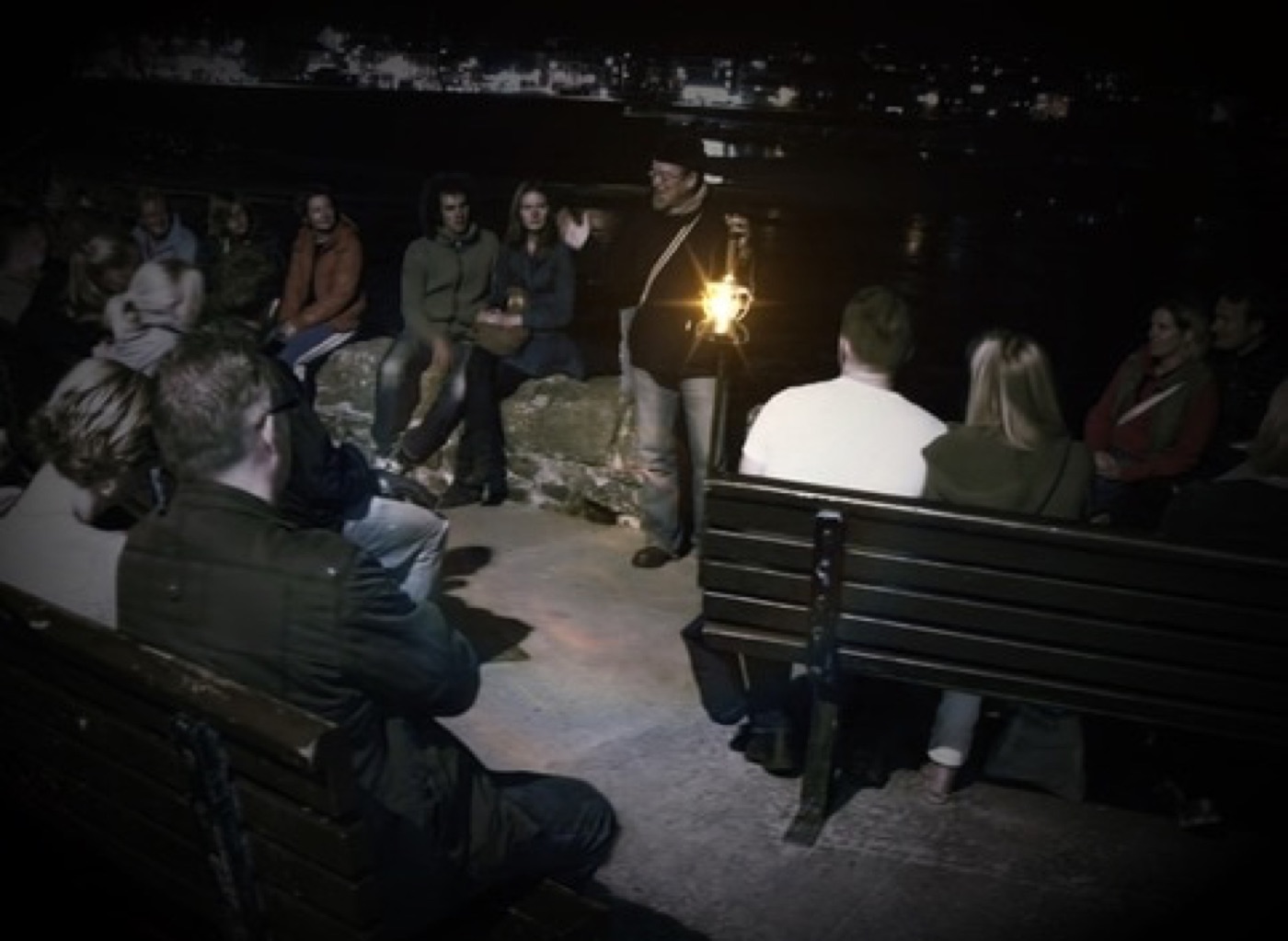 An audience seated in the dark listening to a storyteller holding a lantern