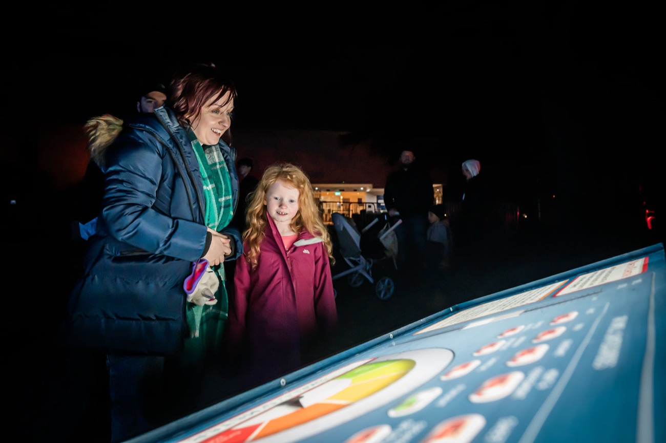 A parent and child smiling at a glowing interactive science exhibit at an evening festival event