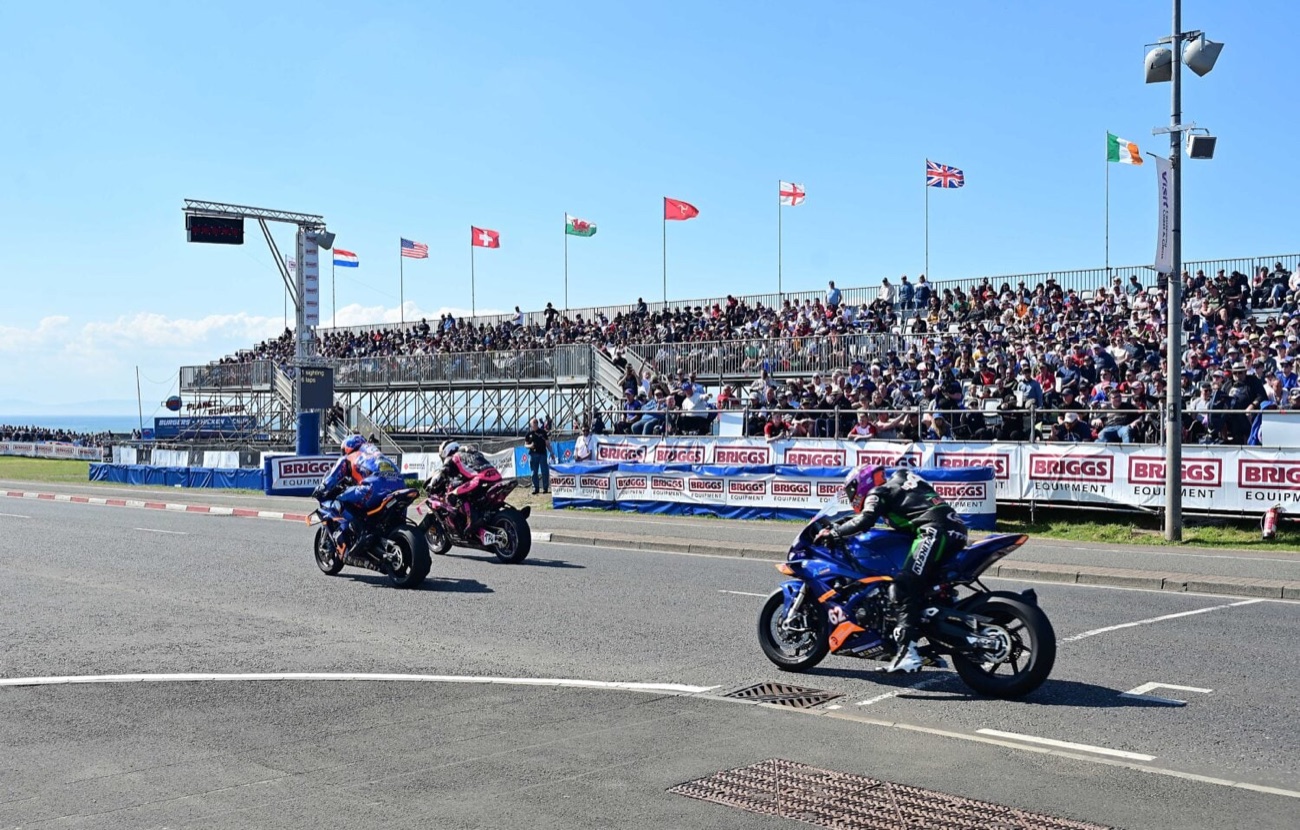 Motorbikes racing past a packed grandstand flying international flags in the sun at the North West 200