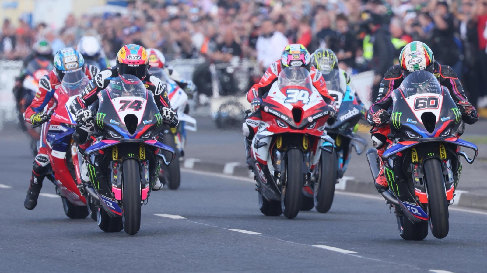 Three superbikes pulling wheelies side by side down the road in front of huge crowds at the North West 200