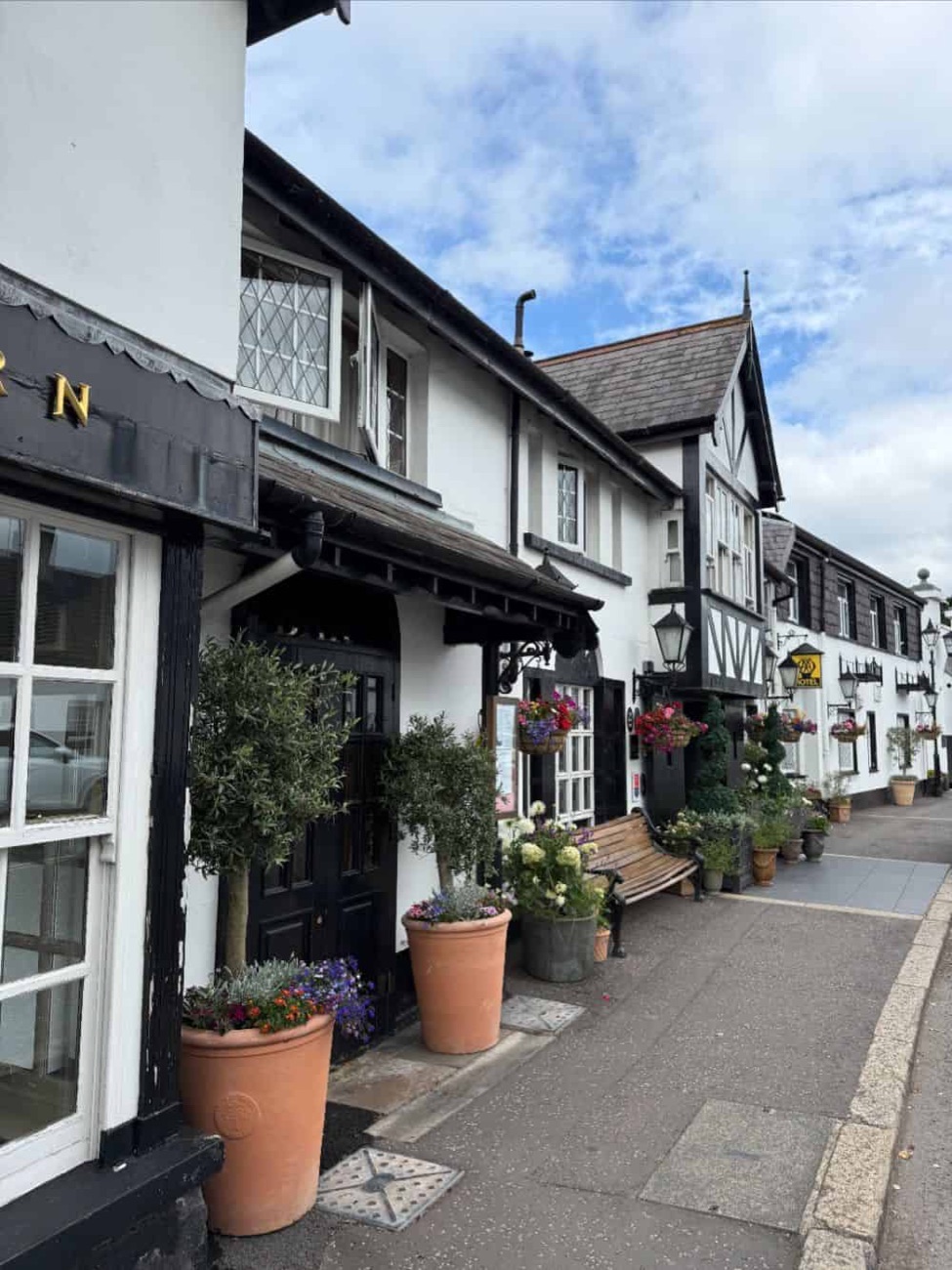 The whitewashed, black-timbered front of The Old Inn with hanging baskets and olive trees.