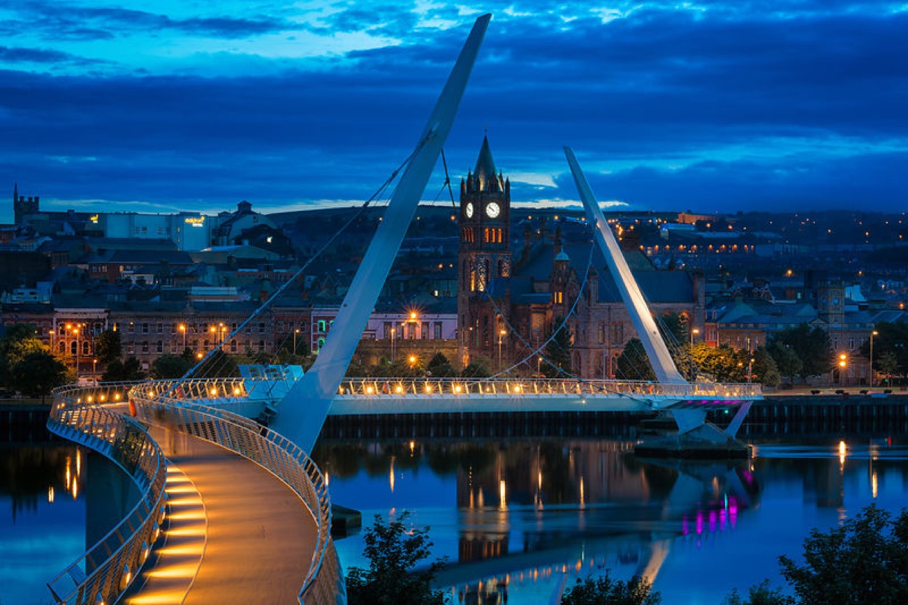 The Peace Bridge at blue hour with the illuminated Guildhall reflected in the calm River Foyle