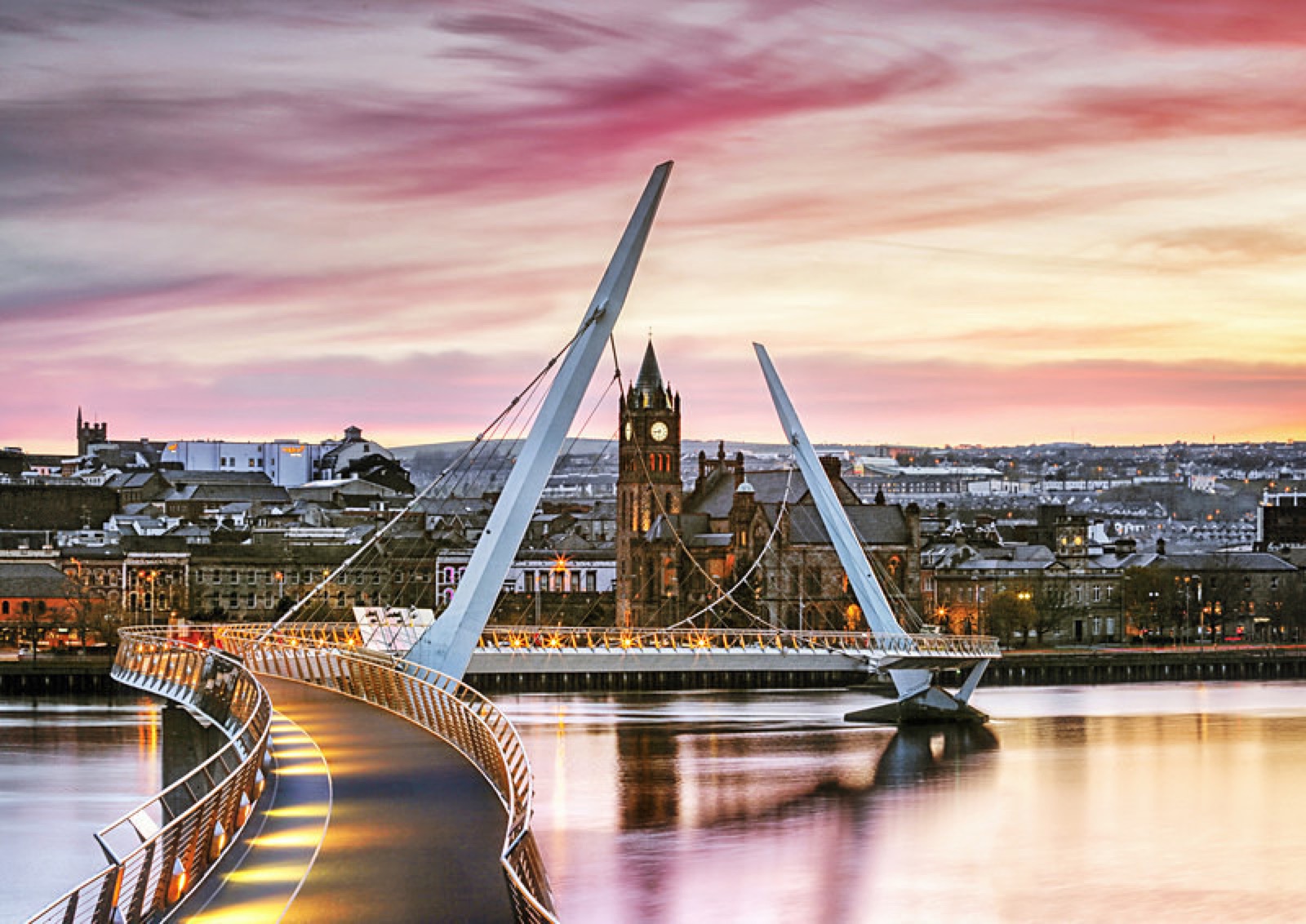 The white cable-stayed Peace Bridge curving across the River Foyle at pink dusk with the Guildhall and Derry city lights behind