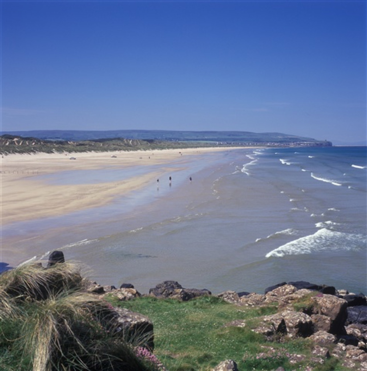 The wide golden beach and rolling surf at Portstewart Strand under a clear blue sky, with the headland stretching into the distance