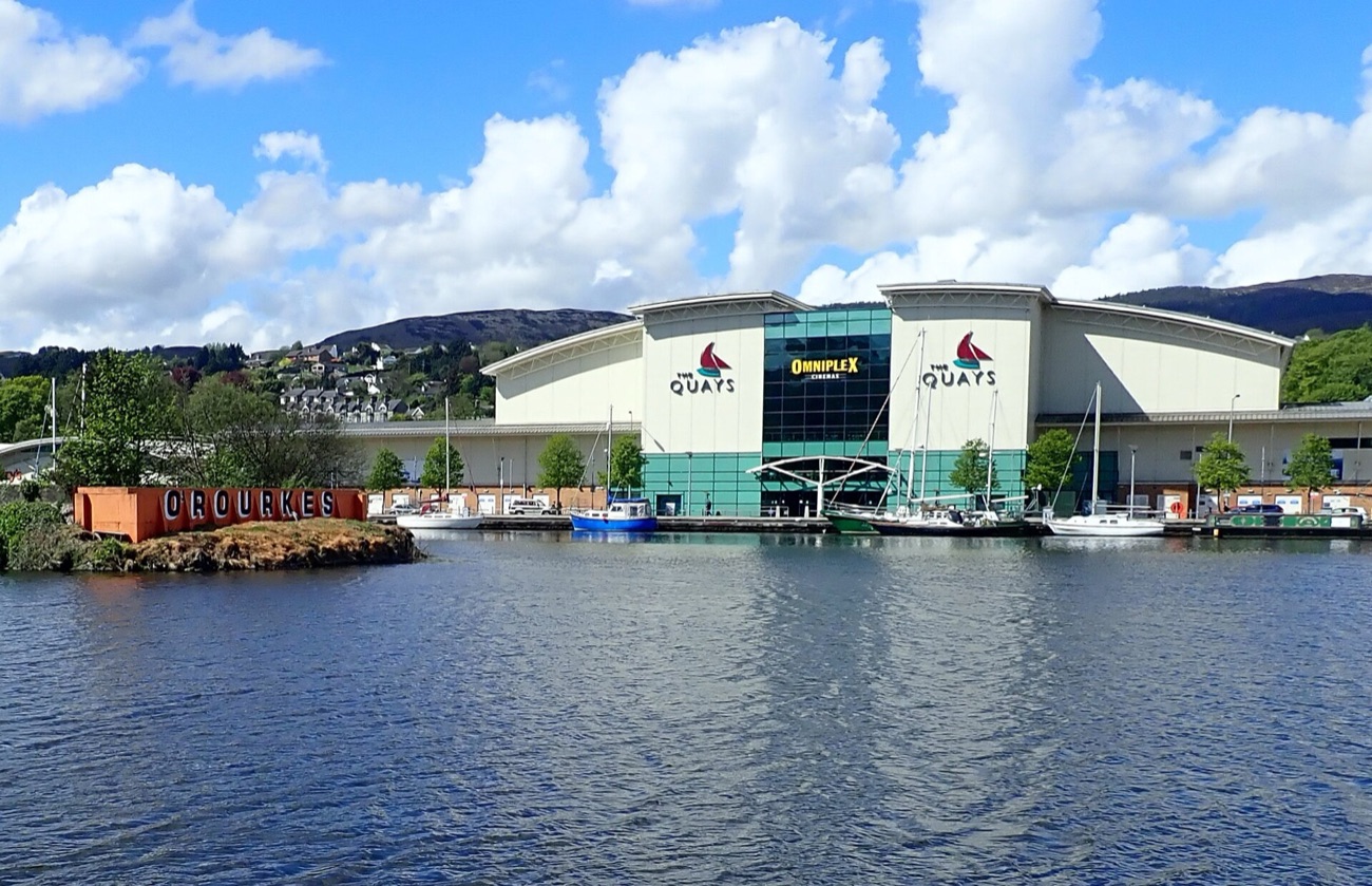 The Quays Shopping Centre and its sail logo seen across the water of the Albert Basin, with boats moored in front and the Mourne Mountains behind