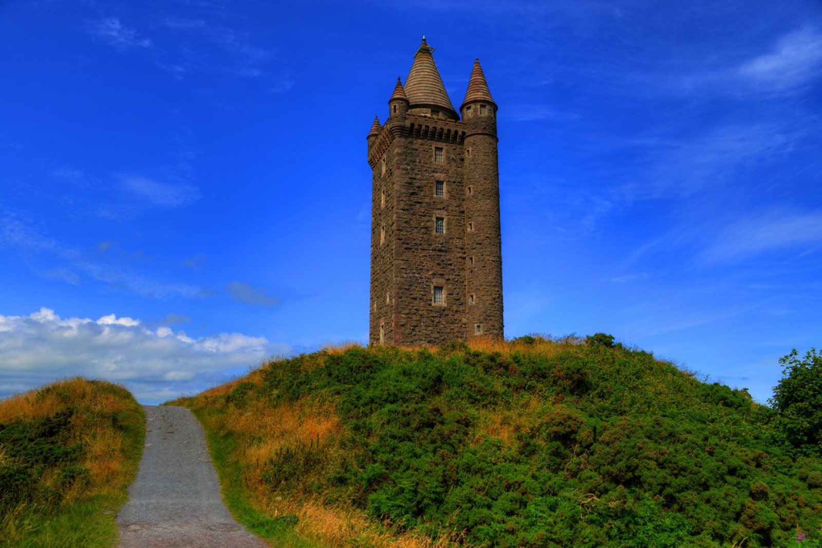 Scrabo Tower, a tall stone tower on its green hilltop with a path leading up under a blue sky