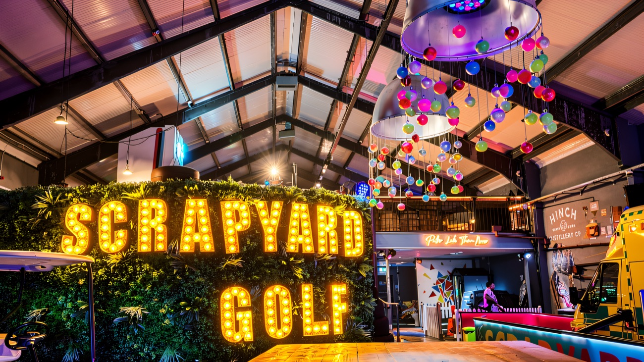 The bulb-lit Scrapyard Golf sign on a foliage wall under hanging disco balls