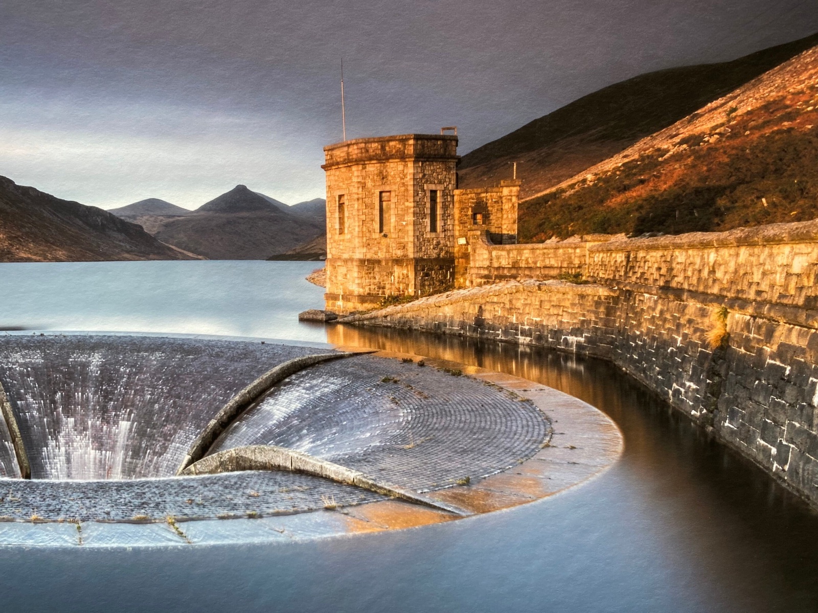 The Silent Valley dam with its stone tower and curved overflow spillway, the Mourne Mountains rising behind in golden light