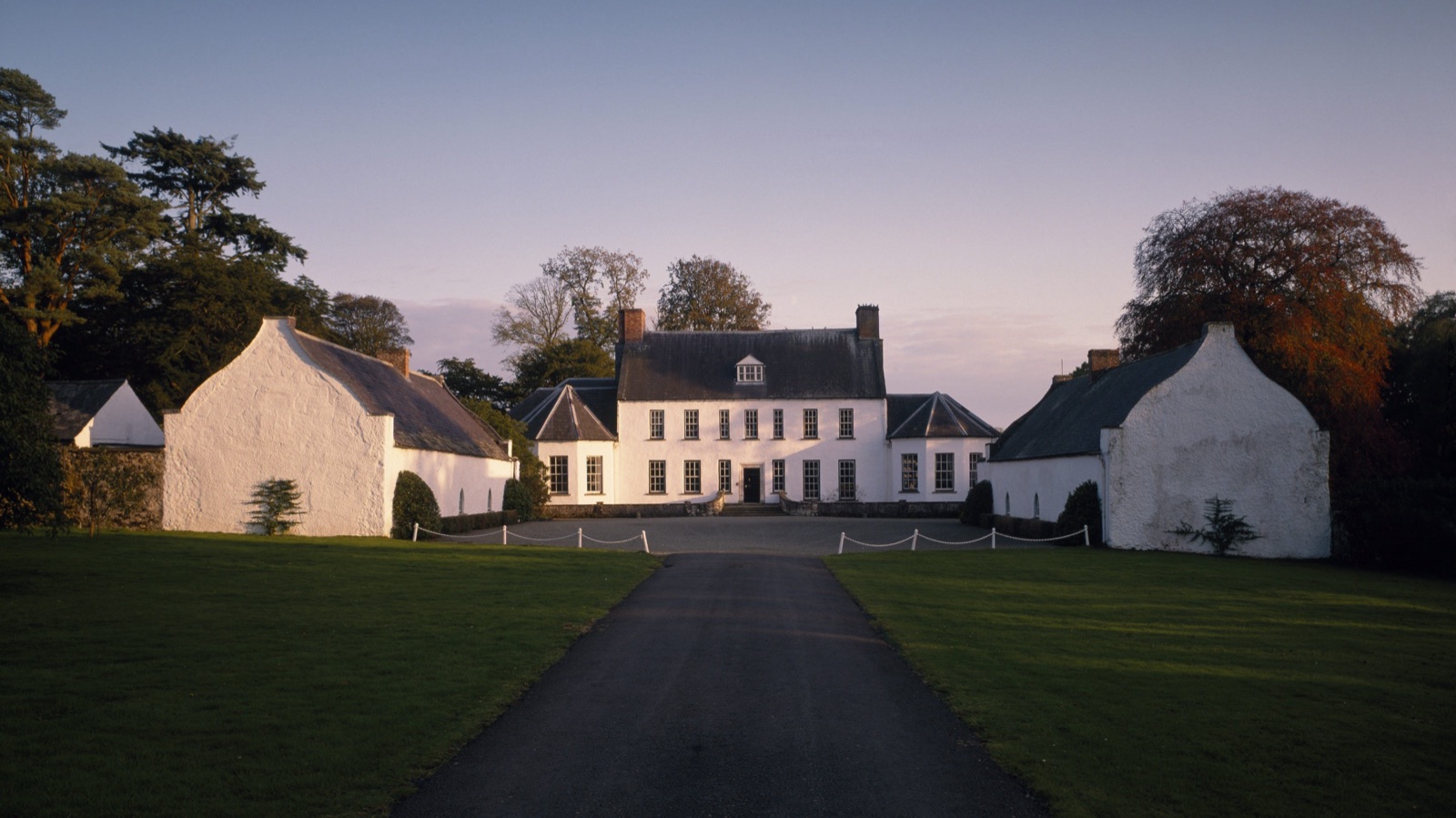 The whitewashed Springhill House at dusk, flanked by two white outbuildings, with a long straight driveway across a green lawn