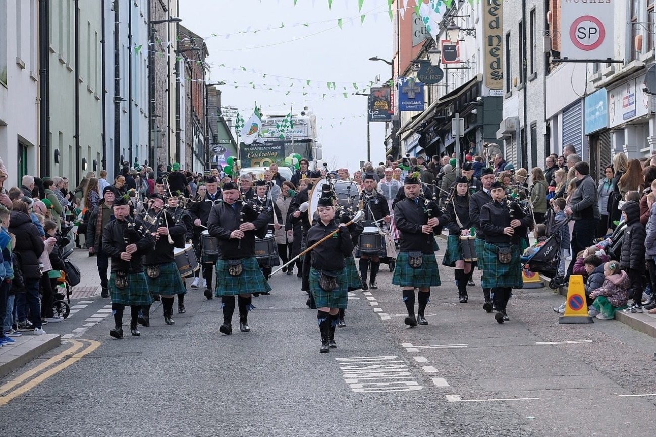 A pipe band in green tartan kilts marching down a Northern Ireland town street on St Patrick's Day