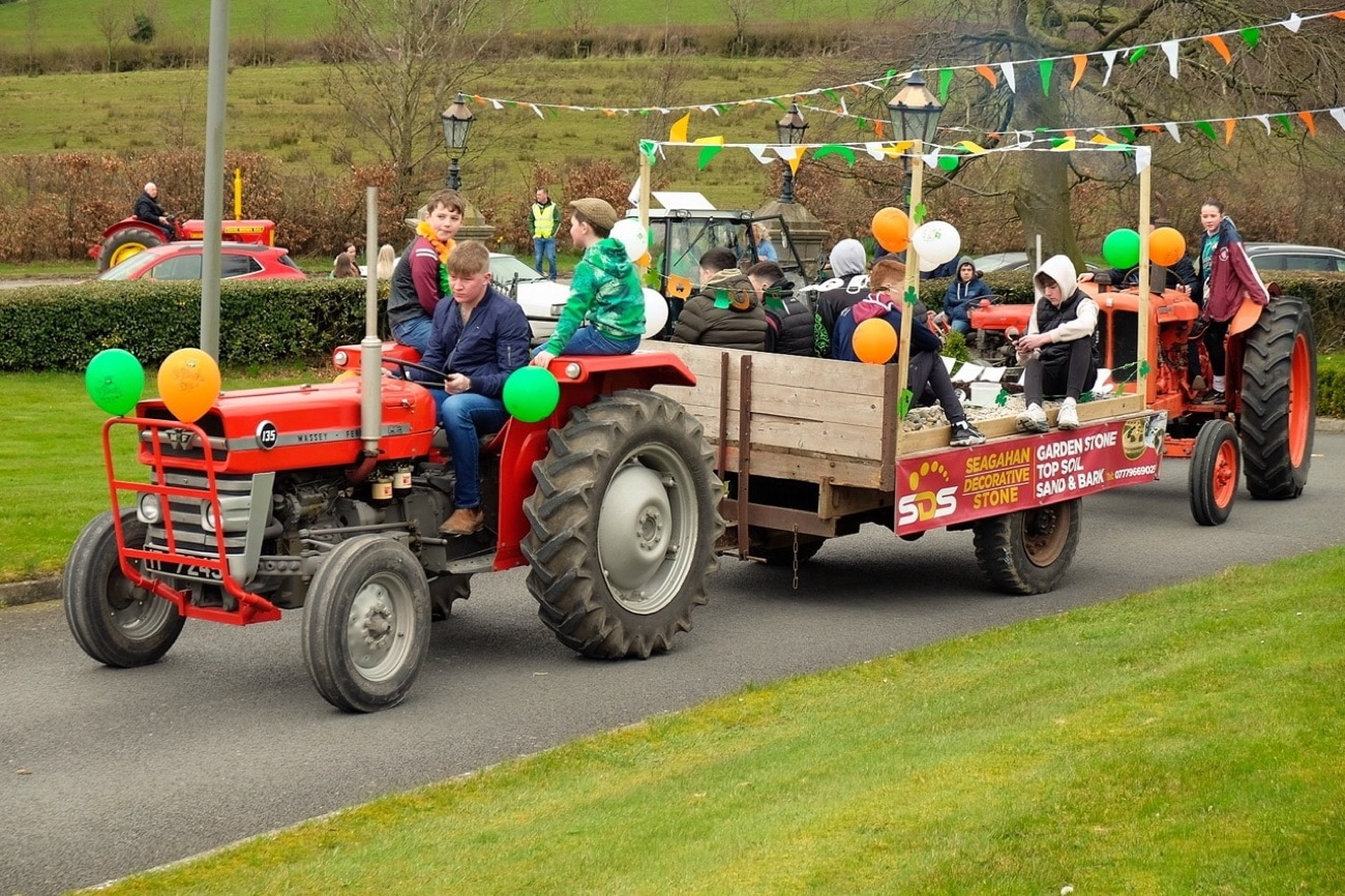 A vintage tractor pulling a decorated trailer float with green and orange balloons in a rural St Patrick's Day parade