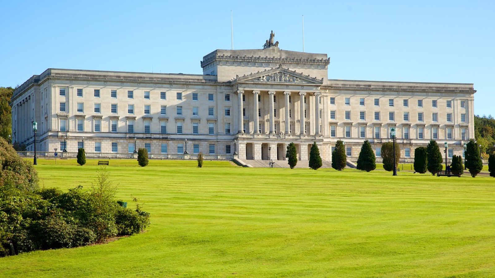 The grand white classical Parliament Buildings at Stormont across a wide green lawn under a blue sky