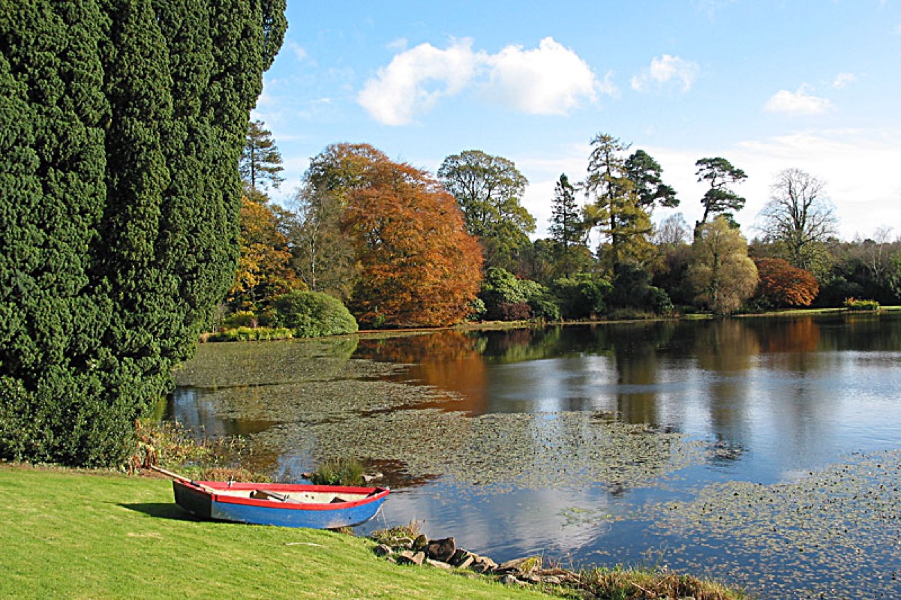 The tree-fringed lough at Tempo Manor with a rowing boat drawn up on the grass and autumn colour reflected in the still water