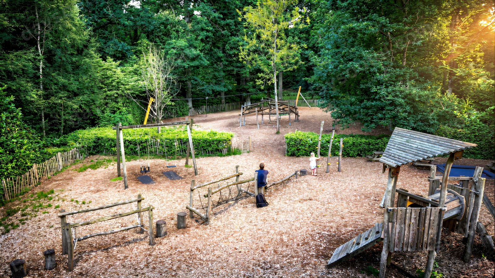 A natural-timber adventure playground among the trees, with climbing frames and balance beams on bark chip.