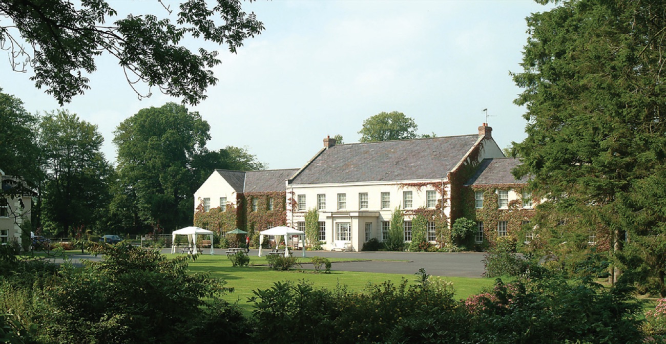 Tullylagan Country House seen across a wide lawn framed by mature trees, with parasols set out on the grass