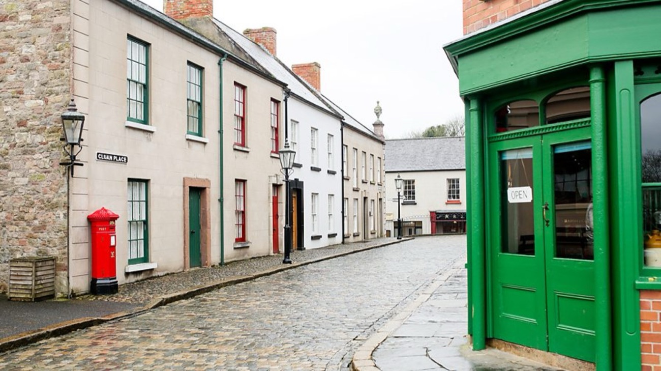 A long low whitewashed thatched cottage with green trim beside a cobbled lane