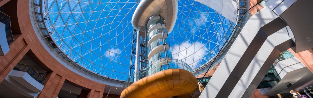 Looking straight up through the atrium of Victoria Square at the glass dome, with the lift tower and viewing pod climbing towards blue sky