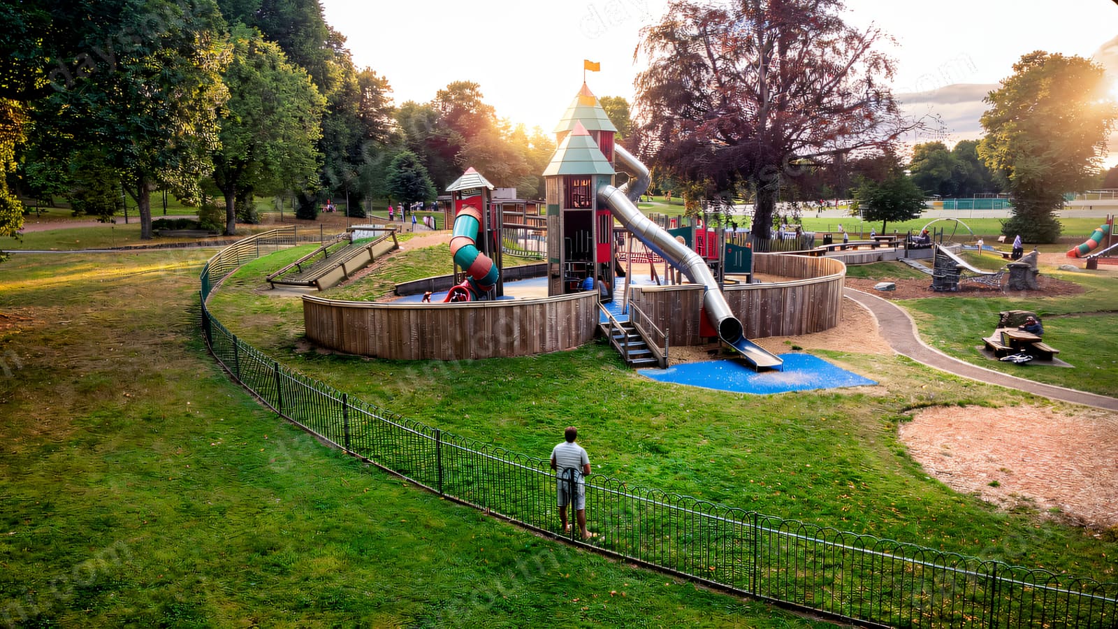 A timber play castle on a raised mound with a long slide, ringed by parkland and old trees.